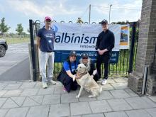 Jay Self on the left with Helena Lee and Kristina Venning crouching down around the guide dog, with Dan Osborne standing on the right, all in front of an Albinism Fellowship banner 