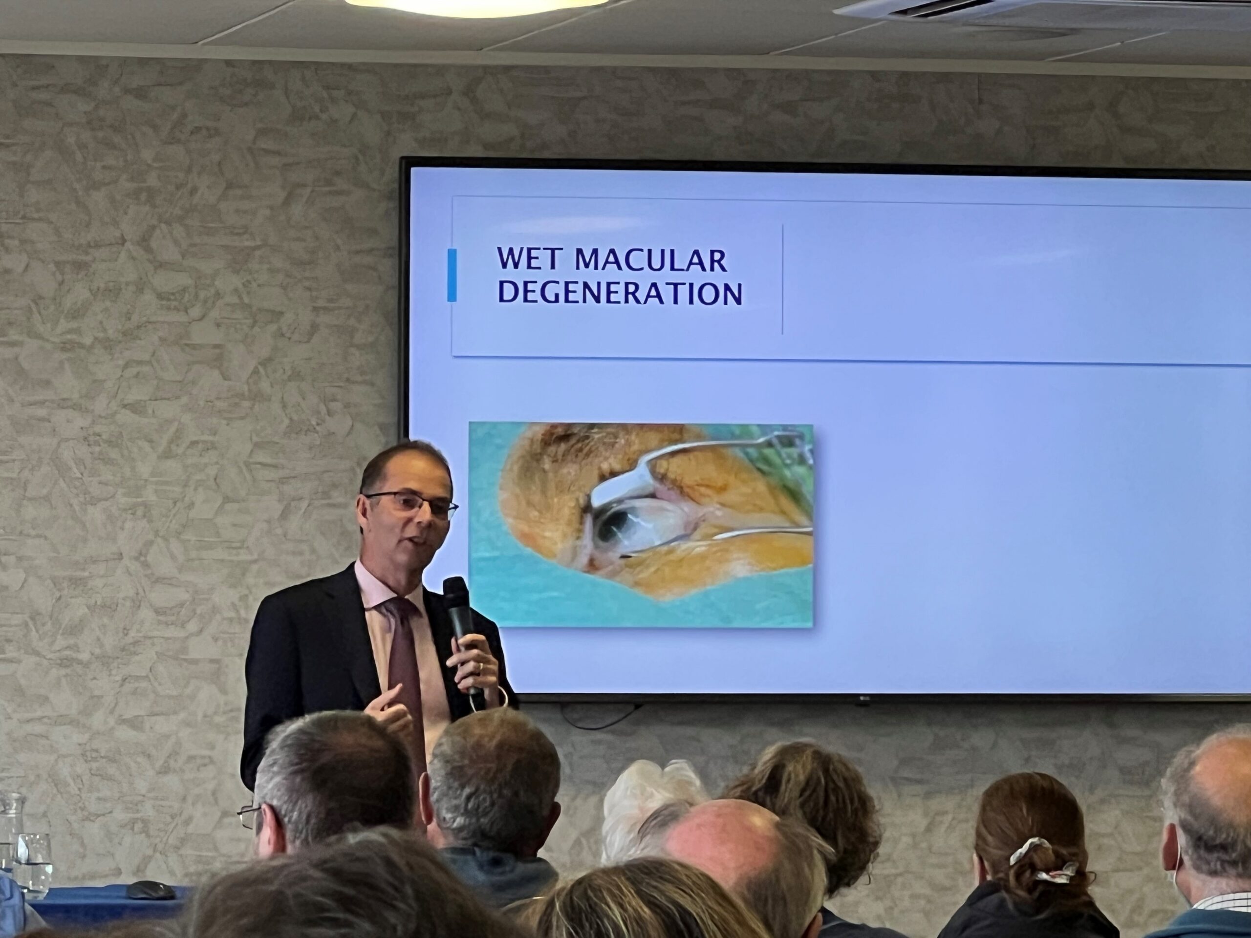 Research into AMD continues Prof Lotery speaking about Wet Macular Degeneration - screen behind him with picture of an eye having injections