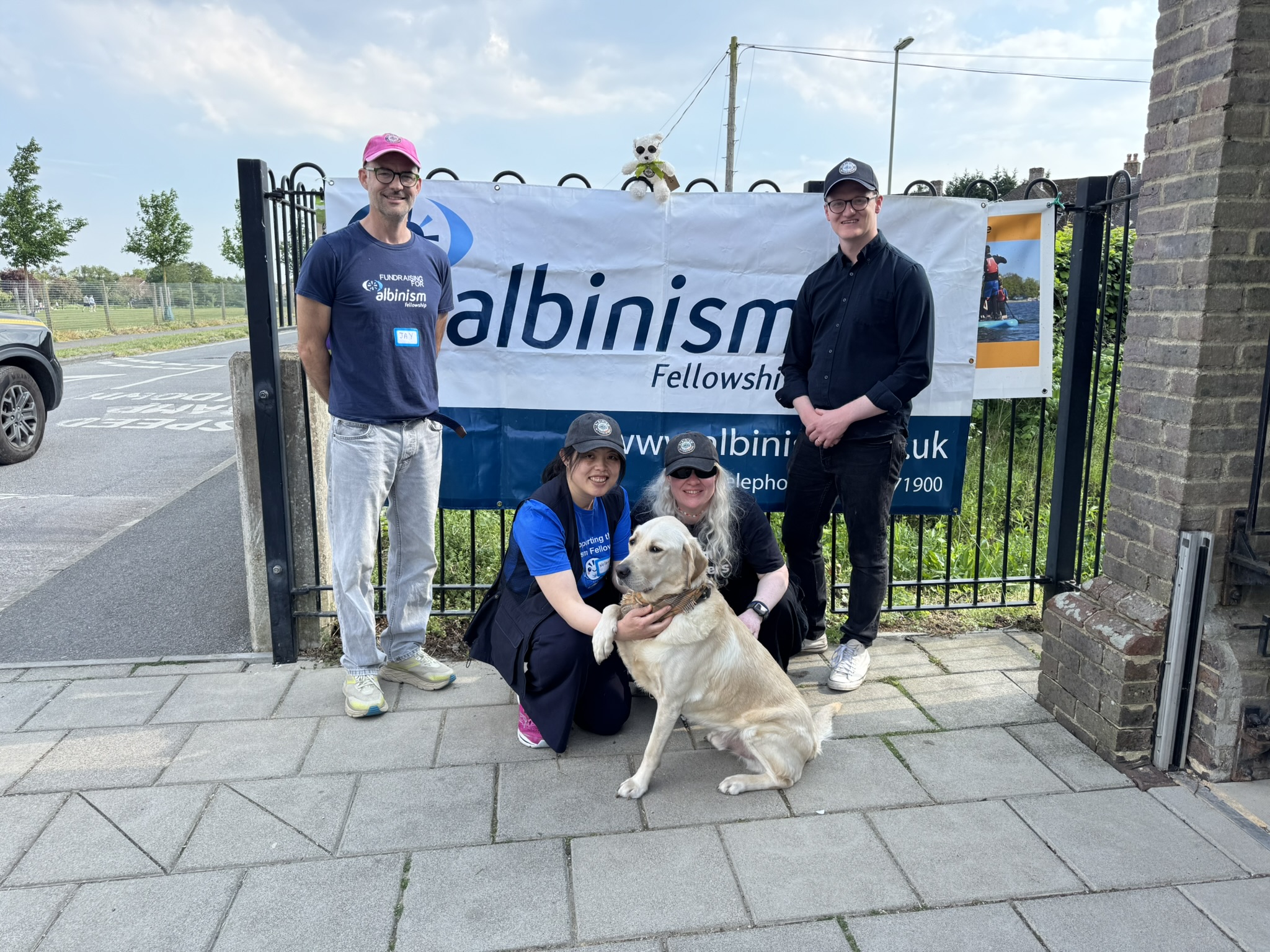 Jay Self on the left with Helena Lee and Kristina Venning crouching down around the guide dog, with Dan Osborne standing on the right, all in front of an Albinism Fellowship banner 