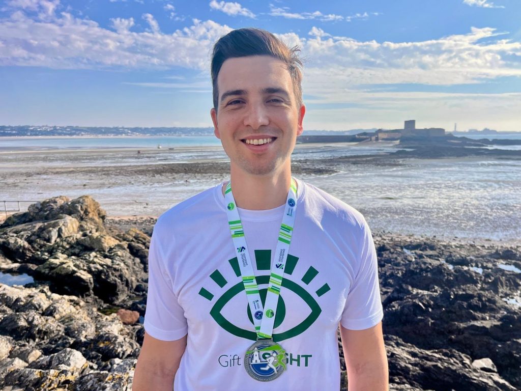 Alex Tanner smiling wearing a Gift of Sight t-shirt with the coastline of Jersey behind him